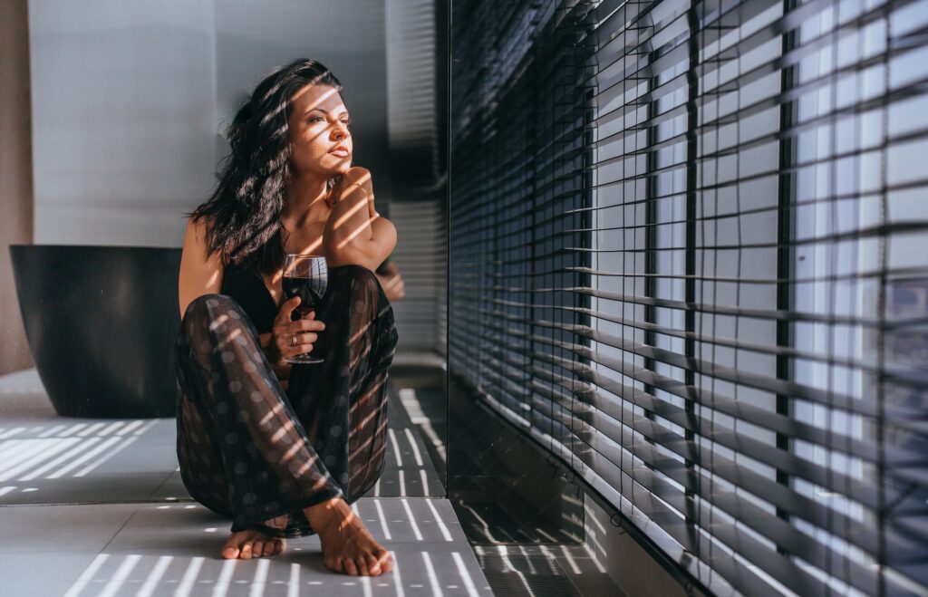 Contemplative woman with glass of wine sitting by window, bathed in sunlight filtered through blinds